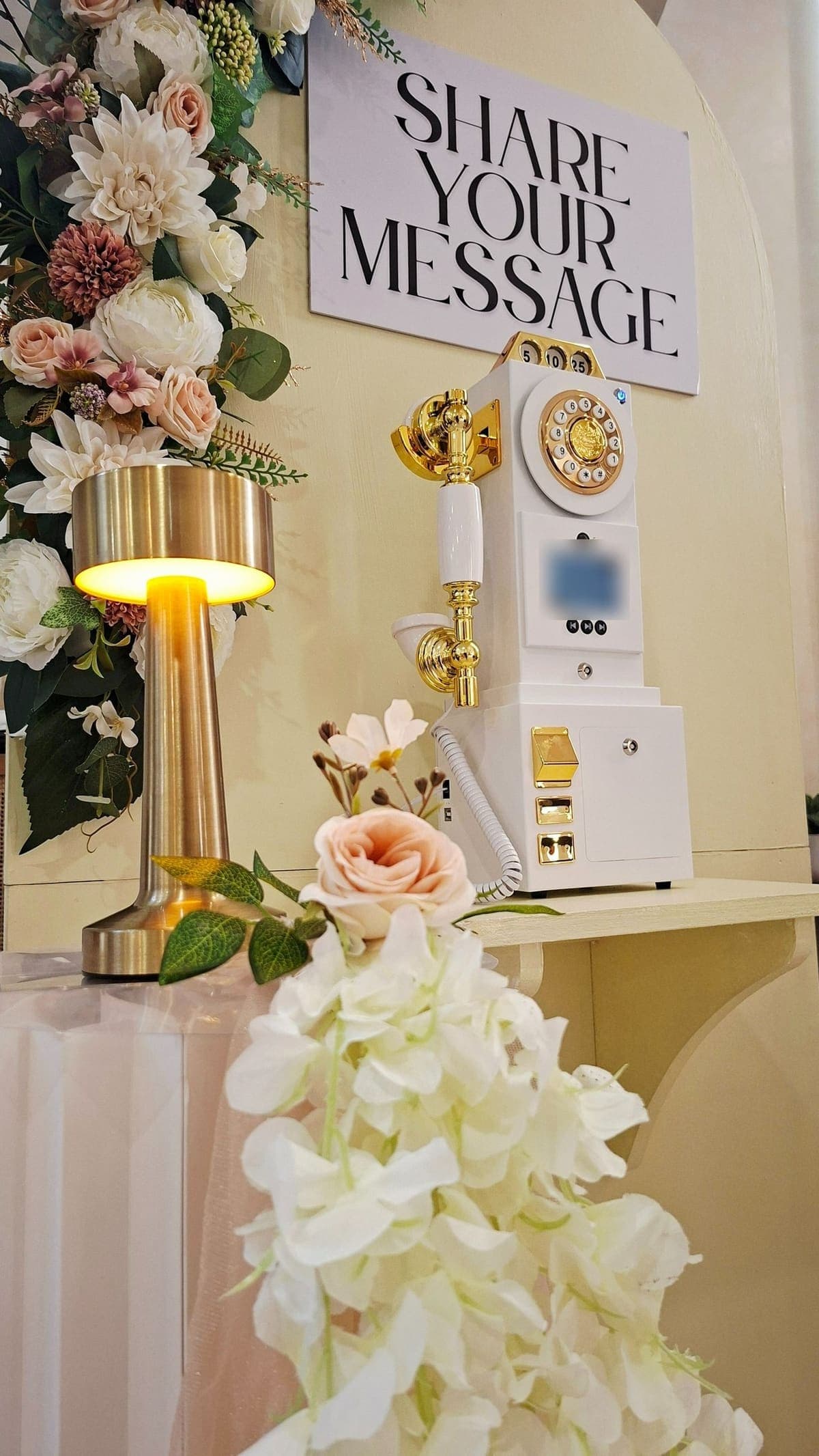 A white-and-gold vintage payphone under a “Share your message” sign, with a brass lamp and blush and ivory flowers